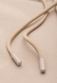 Cordons beige tressés avec embouts en plastique reposant sur un tissu beige texturé.