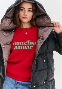 Czarna puchowa kurtka z błyszczącym brązowym wnętrzem, z srebrnymi elementami. Nosi ją osoba w czerwonym swetrze z zielonym napisem "mucho amor".