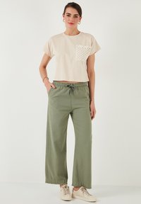 Beige, verkürztes T-Shirt mit strukturiertem Taschen-Design, kombiniert mit lockeren, olivgrünen Hosen mit elastischem Bund und fransigem Saum, getragen mit beige Sneakern.