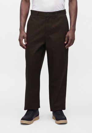 Homme portant un pantalon marron foncé droit, des baskets noires avec des semelles beige, et une chemise blanche sur un fond clair.