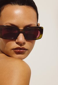 Lunettes de soleil rectangulaires marron avec des verres violets foncés. La monture présente un design massif et une finition brillante sur un fond neutre clair.