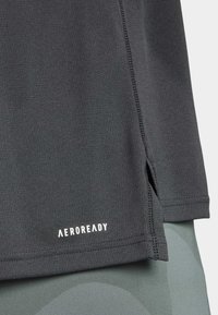 Dunkelgraues Sportshirt aus strukturiertem Material. Verfügt über ein Logo am Saum und einen Seitenschlitz für Belüftung, mit sichtbaren Nähdetails.