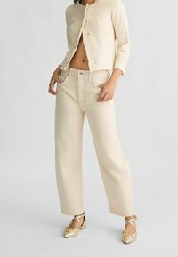 Chaqueta de lino beige con bordes deshilachados, combinada con pantalones de pierna ancha a juego. Los accesorios incluyen zapatos planos dorados con un detalle de lazo.