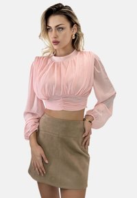 Helles rosa, tailliertes Top mit plissierten Details und langen, puffigen Ärmeln, kombiniert mit einem Minirock aus braunem Wildleder. Das Outfit wird mit einem Armband ergänzt.