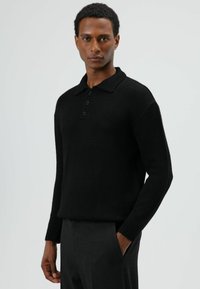 Zwarte gebreide trui met een kraag, een vijfknopenbies en geribbelde manchetten. De stof oogt zacht en met textuur.