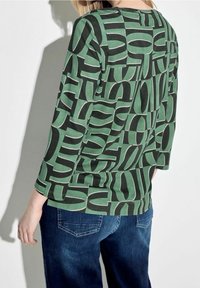 Maglietta verde in cotone a maniche lunghe con un audace motivo astratto nero. Presenta maniche morbide e un orlo curvo, abbinata a jeans blu.