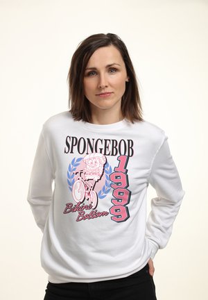Femme portant un sweat blanc avec des illustrations roses et bleues de SpongeBob, le texte "Bikini Bottom" et "1999" à l'avant, debout devant un fond uni.