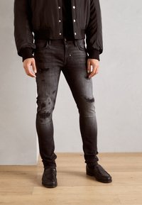 Jeans de mezclilla negros con un corte ajustado, detalles deslavados y parches desgastados. Combinados con una chaqueta bomber negra y zapatos negros.