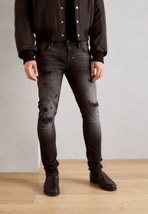 Jeans de mezclilla negros con un corte ajustado, detalles deslavados y parches desgastados. Combinados con una chaqueta bomber negra y zapatos negros.