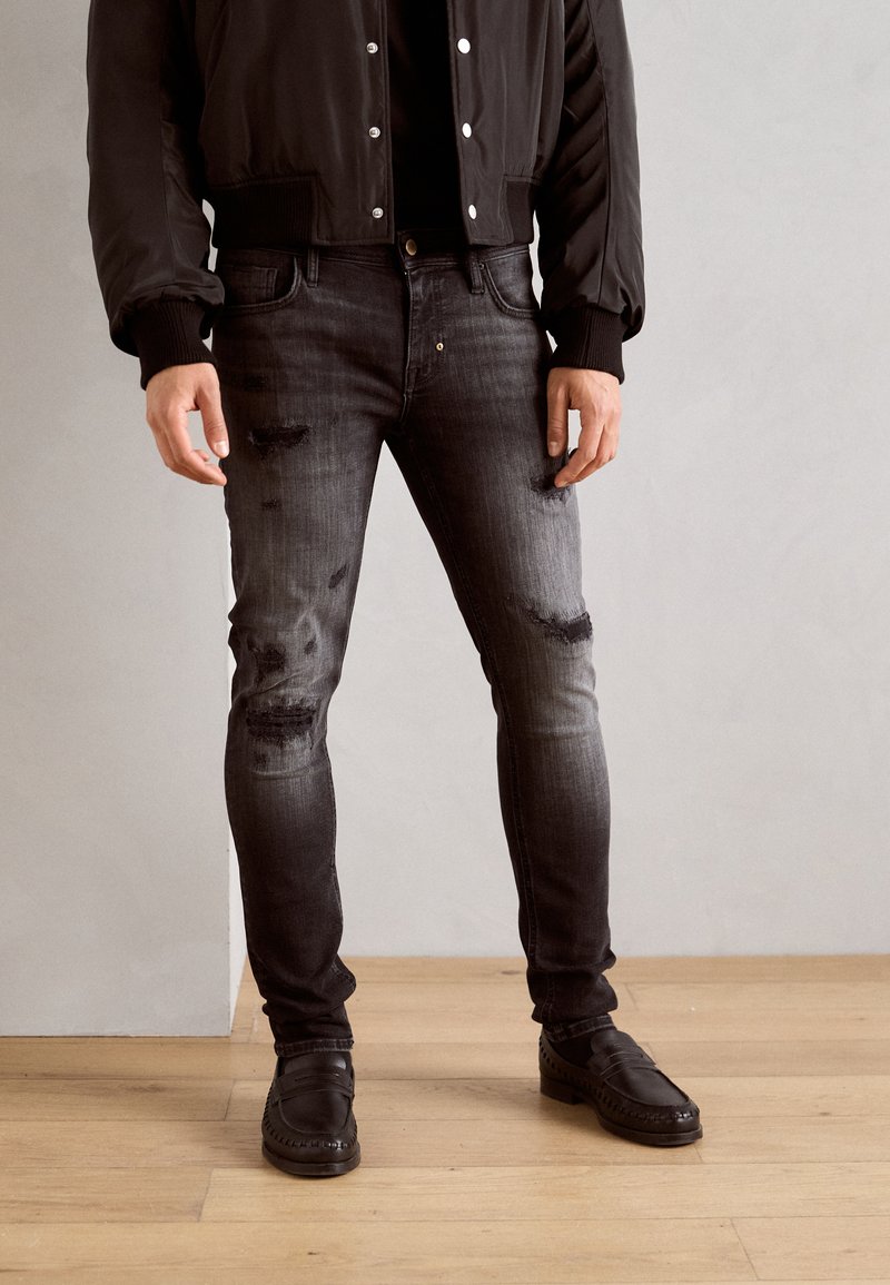 Jeans de mezclilla negros con un corte ajustado, detalles deslavados y parches desgastados. Combinados con una chaqueta bomber negra y zapatos negros.