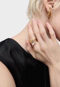 La gioielleria in tono oro include un anello rotondo con una pietra trasparente e una fascia testurizzata, abbinato a un orecchino a cerchio curvo. Sfondo in tessuto nero.