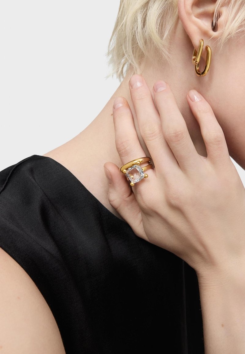 La gioielleria in tono oro include un anello rotondo con una pietra trasparente e una fascia testurizzata, abbinato a un orecchino a cerchio curvo. Sfondo in tessuto nero.