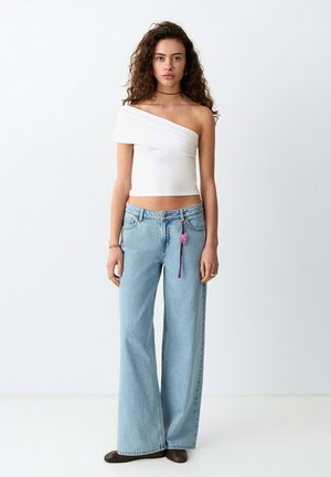 Mlada žena s kovrčavom kosom u bijeloj jednobojnoj majici na jedno rame, širokim plavim trapericama visokog struka i smeđim cipelama stoji uz bijeli zid.