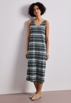 Donna con capelli ricci che indossa un vestito midi senza maniche con motivo a zigzag e scarpe slip-on beige, in piedi contro un muro semplice.