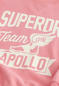 Rosa sweatshirt med stor vit text som säger "SUPERDRY Team JPN" och "APOLLO." Innehåller en sprucken textur och en grafik av en vinge-sko.