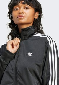 Sort zip-up jakke lavet af glat stof med hvide tre-stribe detaljer på ærmerne og et lille hvidt Adidas-logo på brystet.