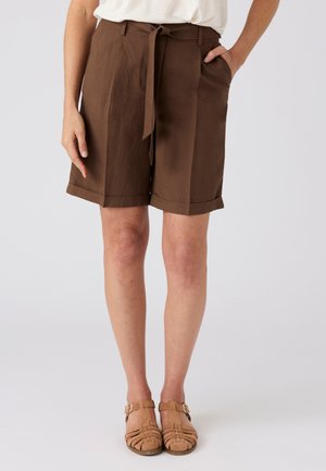 Vrouw draagt bruine op maat gemaakte shorts tot op de knie met ceintuur, wit ingestoken top en beige gesloten sandalen met gespen.