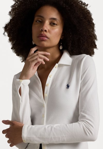 White long-sleeve polo shirt with a collar, featuring a small blue logo, buttoned placket, and unique cutout detail at the cuffs.