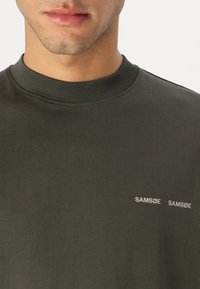 Sweatshirt din bumbac de culoare verde măslin, cu guler rotund cu nervuri. Prezintă un logo din text alb "SAM SøE" pe partea stângă a pieptului. Textură moale, croi lejer.