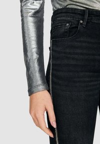 Metallisk silverfärgad långärmad topp i kombination med svarta denimbyxor som har en dragkedjedetalj längs sidan, vilket framhäver en smal passform.