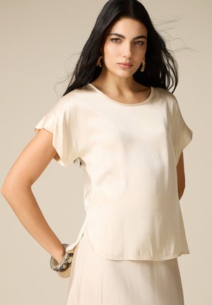 Donna con i capelli lunghi e scuri che indossa una camicetta crema a maniche corte e una gonna abbinata, accessoriata con orecchini dorati e un braccialetto marmorizzato.