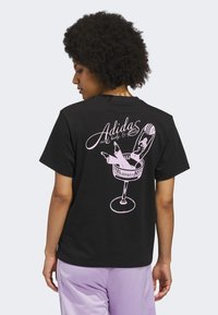 Čierne bavlnené tričko s ružovým grafickým dizajnom ženy v pohári na koktail s textom "Adidas" nad ním. Krátke rukávy, voľný strih.