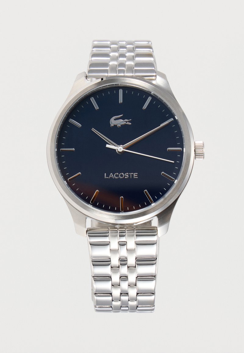 Montre-bracelet en métal argenté avec cadran bleu marine, marqueurs d'heure minimalistes, logo Lacoste, et trois aiguilles indiquant 10:10:18.