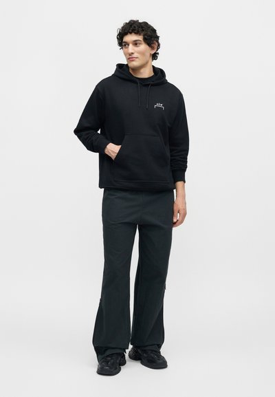 Νέος άνδρας που στέκεται, φοράει μαύρο φούτερ με κουκούλα και λογότυπο, μαύρο παντελόνι με φαρδιά πόδια και μαύρα αθλητικά παπούτσια, κοιτάζει προς το πλάι.
