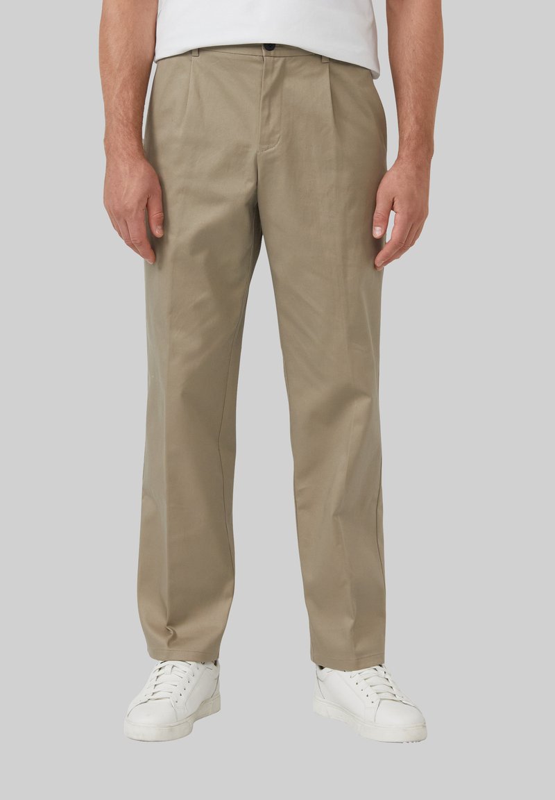 Beige broeken met een ontspannen pasvorm, met een geplooide voorkant, een lichte glans en gedetailleerde stiksels, gecombineerd met witte sneakers op een grijze achtergrond.