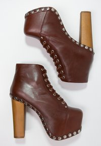 Botas de tobillo de plataforma de cuero marrón con diseño de cordones, acentos de tachuelas plateadas y un tacón de madera gruesa, presentando una textura suave.