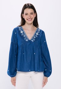 Denimbluse mit V-Ausschnitt, verziert mit floraler Stickerei am Ausschnitt, langen Puffärmeln und einer lockeren Passform. Hellblaue Farbe.
