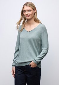 Ljusgrön v-ringad tröja i mjukt tyg, med långa ärmar och figurnära kant. Modellen bär den tillsammans med mörkblå jeans.