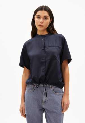 Navy satin blouse with short sleeves, button front, and a drawstring hem. Includes a chest pocket. Paired with light-wash jeans.