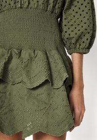 Robe vert olive avec manches bouffantes en oeillets, taille smockée et ourlet brodé festonné, portée sur le torse et le bras d'une personne.