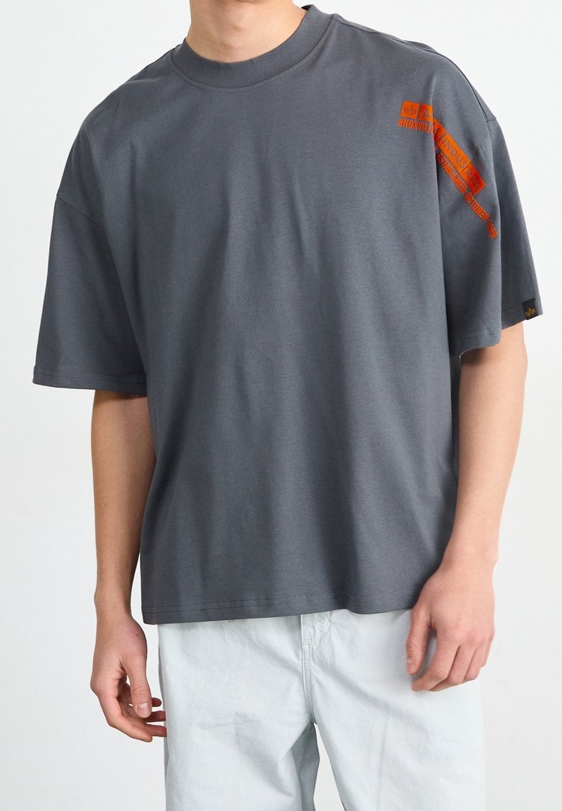 T-shirt en coton gris à manches courtes, col rond, avec un texte graphique orange sur la poitrine et l'épaule. Coupe décontractée, texture lisse.