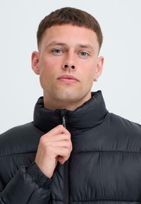Svart pufferjacka med hög krage och dragkedja fram. Har quiltade sektioner och elastiska muddar, vilket ger en snygg och modern design.