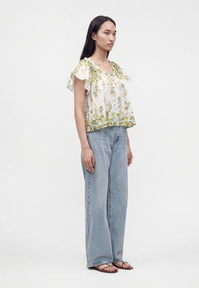Femme portant un chemisier blanc à manches courtes à motif floral, un jean large bleu clair et des sandales marron, debout sur un fond blanc uni.