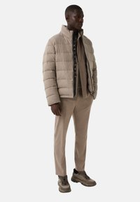 Mand iført en beige pufferjakke, en matchende beige skjorte med knapper, skræddersyede bukser og beige sneakers, der står foran en ensfarvet baggrund.
