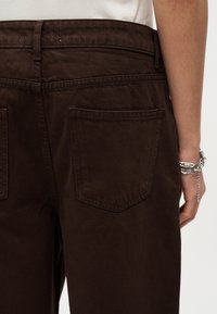 Άτομο που φοράει σκούρο καφέ παντελόνι με πίσω τσέπες, λευκό μπλουζάκι που έχει μπει μέσα στο παντελόνι και ασημένιο βραχιόλι με αλυσίδα στον δεξί καρπό.