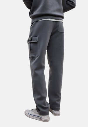 Osoba ubrana w ciemnoszare spodnie dresowe z boczną kieszenią cargo oraz jasnoszare trampki, stojąca tyłem do kamery.