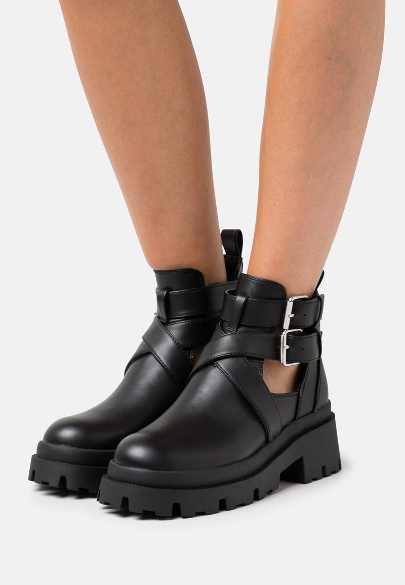 Jambes portant des bottines en cuir noir épaisses avec des semelles épaisses, des brides à double boucle et des côtés découpés sur fond blanc.