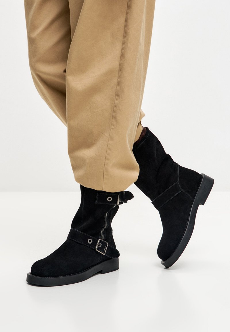 Crne antilop gležnjače s okruglim vrhom, bočnim patentnim zatvaračem, trakom s kopčom i ravnim gumanim potplatom, uparene s bež hlačama.