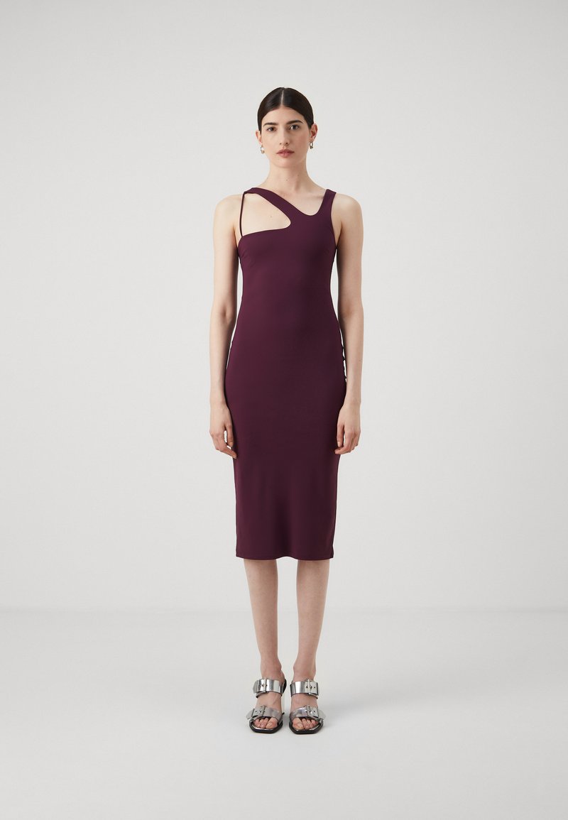 Burgundowa sukienka bodycon z asymetrycznym dekoltem i pojedynczym paskiem, wykonana z gładkiego materiału. Modelka nosi srebrne sandały z metalowymi akcentami.
