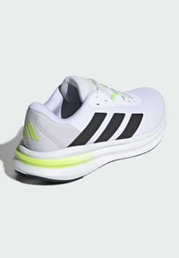 Zapatilla deportiva blanca con parte superior de malla, rayas negras, acentos reflectantes y detalles en verde neón sobre una suela acolchada. Cuenta con cierre de cordones.