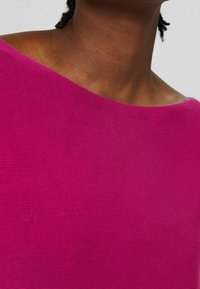 Gros plan sur une personne portant un haut en tricot rose vif à épaules dénudées, montrant le cou, la clavicule et une partie de la coiffure, sur un fond neutre.