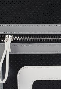 Zwart geperforeerd materiaal met grijze accenten, witte details en een zilveren rits. Beschikt over een bovenritssluiting en zichtbare stiksels.