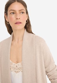 Cardigan beige con colletto a scialle a costine, design aperto e texture morbida. Indossato sopra un top bianco con bordi in pizzo.