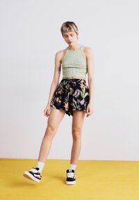 Zöld csíkozott crop top, virágmintás fekete rövidnadrág, megkötős derékpánttal, laza fekete-fehér sportcipő, sárga padlón állva, semleges háttérrel.