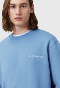 Světlemodrá mikina vyrobená z bavlny, s kulatým výstřihem, oversized střihem a bílým potiskem loga na hrudi. Hladká textura.