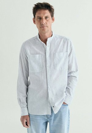 Hombre con pelo corto y castaño que lleva una camisa blanca de manga larga con finas rayas verticales negras y vaqueros azul claro, de pie frente a un fondo liso.